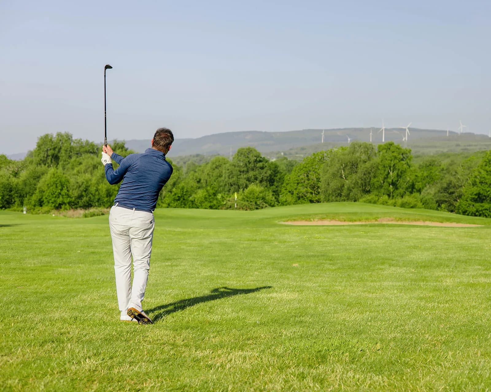 Stuart Wilson at the top of his backswing on an approach to a bunkered green, with wind turbines on the hills behind